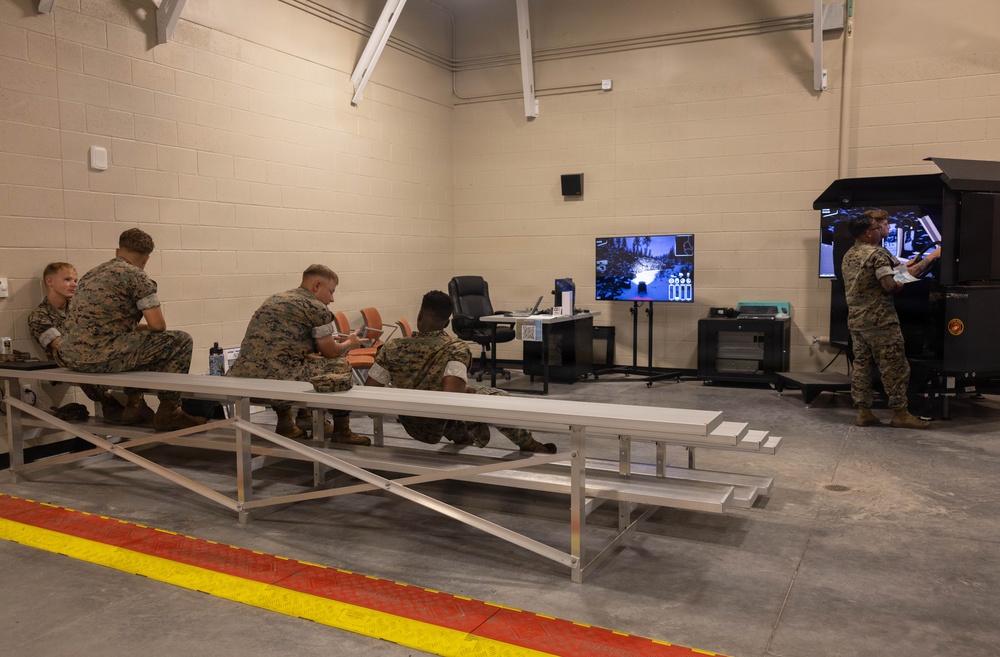 A group of military personnel in camouflage uniforms are gathered in what appears to be a training facility or simulation room. Several individuals are seated on metal bleachers watching a display screen showing what appears to be a tactical simulation, while one person operates a enclosed simulator unit on the right side of the room. The setting is an institutional interior space with concrete block walls and a marked floor.