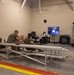 A group of military personnel in camouflage uniforms are gathered in what appears to be a training facility or simulation room. Several individuals are seated on metal bleachers watching a display screen showing what appears to be a tactical simulation, while one person operates a enclosed simulator unit on the right side of the room. The setting is an institutional interior space with concrete block walls and a marked floor.