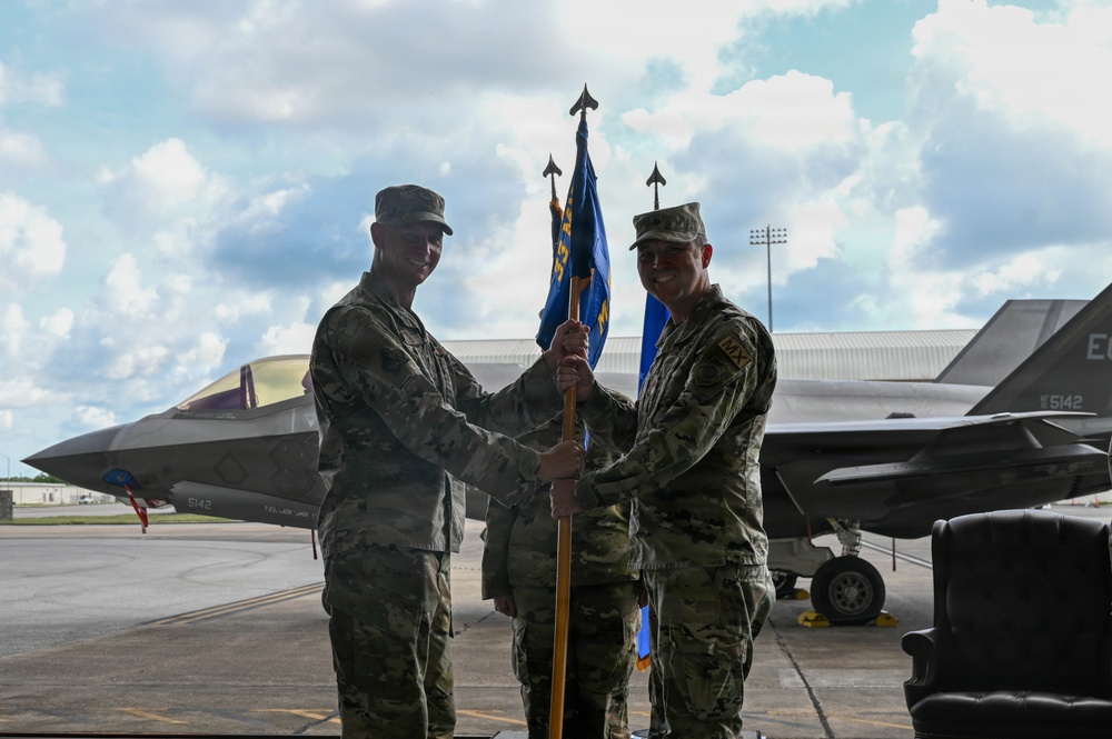 Two U.S. Air Force personnel in MultiCam/OCP camouflage uniforms are conducting a guidon/pennant passing ceremony on a flight line, consistent with a change of command or activation ceremony. Behind them are at least two F-35 Lightning II stealth fighter jets bearing tail number 5142. A third uniformed individual stands in the background.