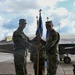 Two U.S. Air Force personnel in MultiCam/OCP camouflage uniforms are conducting a guidon/pennant passing ceremony on a flight line, consistent with a change of command or activation ceremony. Behind them are at least two F-35 Lightning II stealth fighter jets bearing tail number 5142. A third uniformed individual stands in the background.