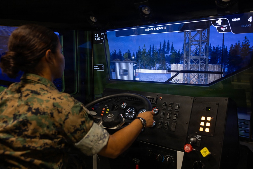 A U.S. Marine in digital camouflage (MARPAT) uniform is operating a military vehicle driving simulator. The simulator displays a virtual environment showing a military installation or base with trees, buildings, and a tower. The screen indicates 'END OF EXERCISE' with telemetry data including speed (24 MPH), RPM (1,652), and gear (4).