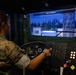 A U.S. Marine in digital camouflage (MARPAT) uniform is operating a military vehicle driving simulator. The simulator displays a virtual environment showing a military installation or base with trees, buildings, and a tower. The screen indicates 'END OF EXERCISE' with telemetry data including speed (24 MPH), RPM (1,652), and gear (4).
