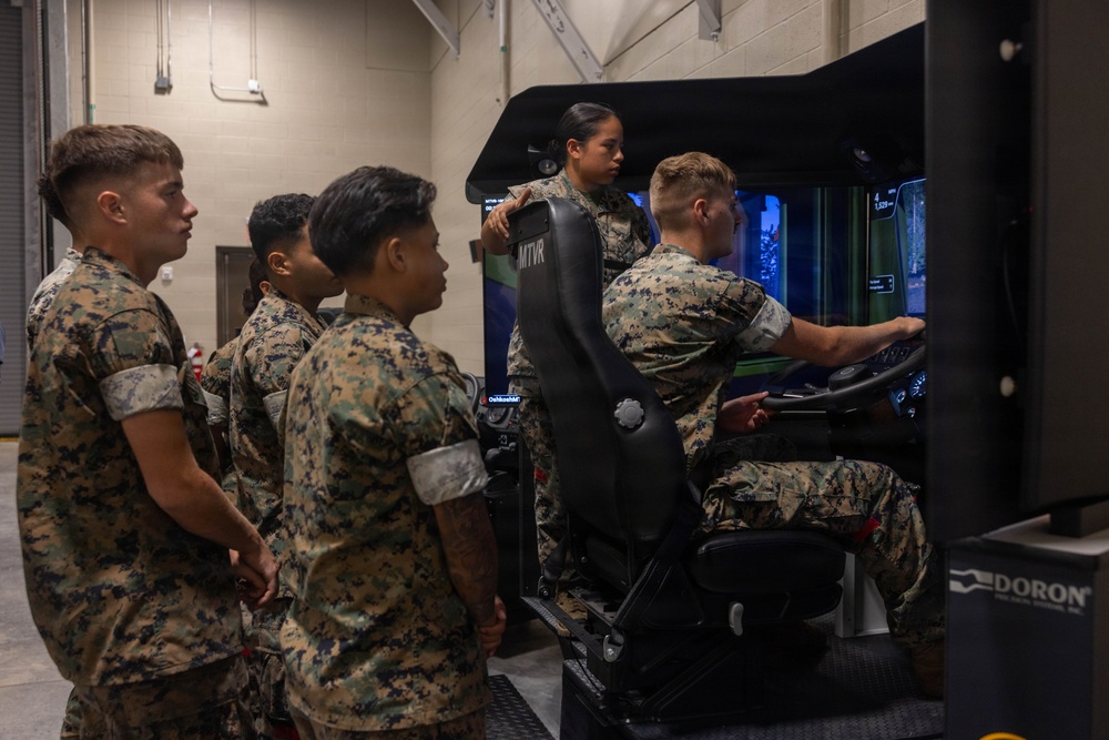 A group of military personnel in MARPAT camouflage uniforms are gathered around a vehicle driving simulator. One service member is actively operating the simulator while others observe, with an instructor standing behind overseeing the training session. The simulator is identified as an MTVR (Medium Tactical Vehicle Replacement) trainer manufactured by Doron Precision Systems.