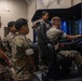 A group of military personnel in MARPAT camouflage uniforms are gathered around a vehicle driving simulator. One service member is actively operating the simulator while others observe, with an instructor standing behind overseeing the training session. The simulator is identified as an MTVR (Medium Tactical Vehicle Replacement) trainer manufactured by Doron Precision Systems.