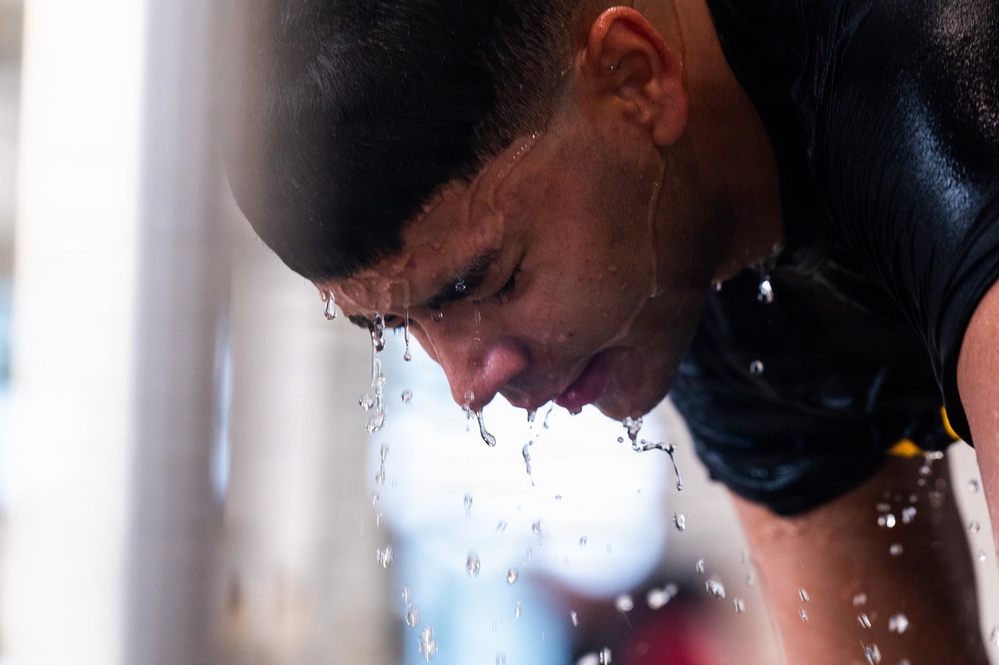 A close-up photograph of a young man leaning forward with water cascading down his face in dramatic streams and droplets. He is wearing a dark athletic shirt, suggesting a sports or physical training context. The image captures a moment of intense physical exertion or cooling down after activity.
