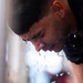 A close-up photograph of a young man leaning forward with water cascading down his face in dramatic streams and droplets. He is wearing a dark athletic shirt, suggesting a sports or physical training context. The image captures a moment of intense physical exertion or cooling down after activity.