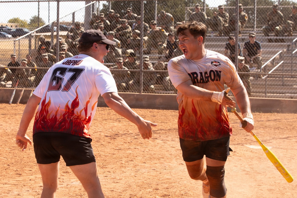 Two players wearing matching white and flame-patterned 'Dragon' team jerseys celebrate on a dirt softball/baseball field. The player on the right, holding a yellow bat, appears to be shouting in excitement while the player wearing number 67 reaches toward him. A large crowd of what appears to be military personnel in uniform watches from behind a chain-link fence in the background.