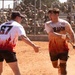 Two players wearing matching white and flame-patterned 'Dragon' team jerseys celebrate on a dirt softball/baseball field. The player on the right, holding a yellow bat, appears to be shouting in excitement while the player wearing number 67 reaches toward him. A large crowd of what appears to be military personnel in uniform watches from behind a chain-link fence in the background.