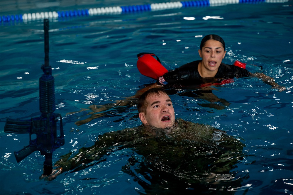 Two individuals are in an indoor swimming pool during what appears to be a military or law enforcement water survival training exercise. A male trainee in camouflage uniform is swimming while holding a rifle above water, with a female instructor or fellow trainee wearing red flotation arm bands positioned behind him. The setting is a standard lap pool with blue lane dividers visible in the background.