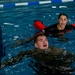 Two individuals are in an indoor swimming pool during what appears to be a military or law enforcement water survival training exercise. A male trainee in camouflage uniform is swimming while holding a rifle above water, with a female instructor or fellow trainee wearing red flotation arm bands positioned behind him. The setting is a standard lap pool with blue lane dividers visible in the background.
