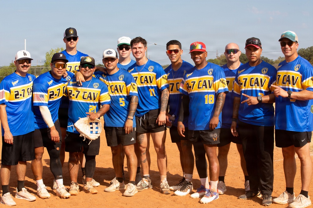A group of approximately 13 men pose together on a dirt baseball/softball field wearing matching blue, gold, and white jerseys labeled '2-7 CAV'. The team appears to be a military unit softball team, as '2-7 CAV' likely refers to the 2nd Battalion, 7th Cavalry Regiment. The photo is a casual team portrait taken outdoors in sunny conditions.