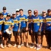 A group of approximately 13 men pose together on a dirt baseball/softball field wearing matching blue, gold, and white jerseys labeled '2-7 CAV'. The team appears to be a military unit softball team, as '2-7 CAV' likely refers to the 2nd Battalion, 7th Cavalry Regiment. The photo is a casual team portrait taken outdoors in sunny conditions.