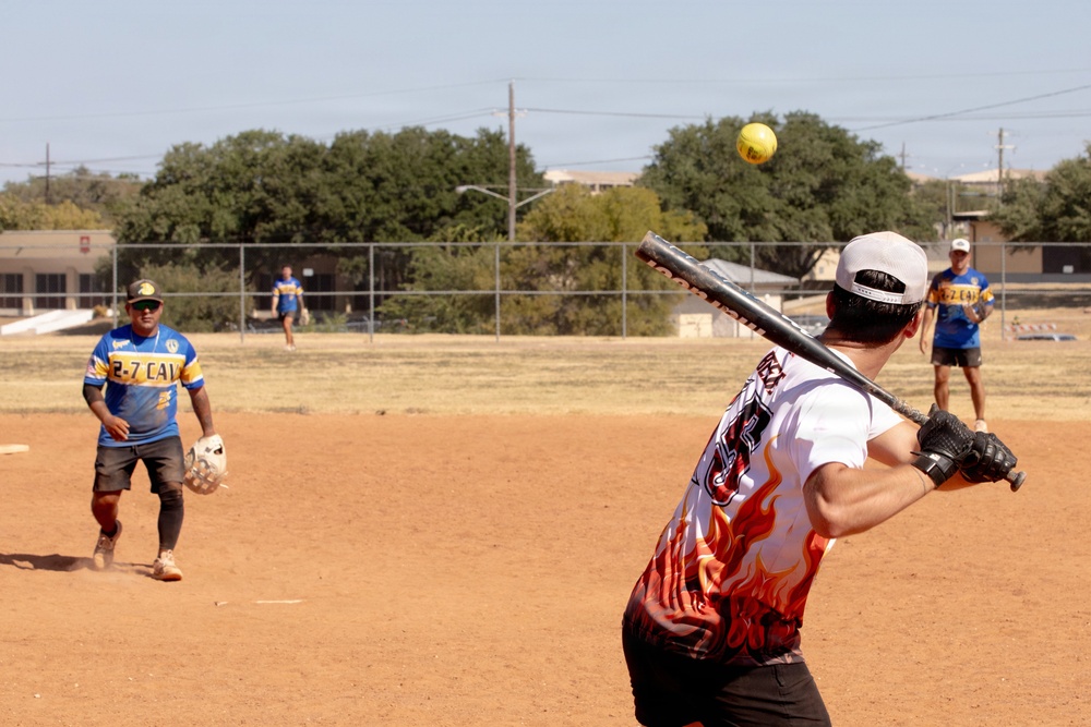 A recreational softball game is in progress on a dirt field under clear sunny skies. A batter wearing a white flame-patterned jersey and backwards cap is mid-swing at a yellow softball. Fielders wearing blue and gold '2-7 CAV' jerseys are positioned around the infield.