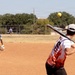 A recreational softball game is in progress on a dirt field under clear sunny skies. A batter wearing a white flame-patterned jersey and backwards cap is mid-swing at a yellow softball. Fielders wearing blue and gold '2-7 CAV' jerseys are positioned around the infield.