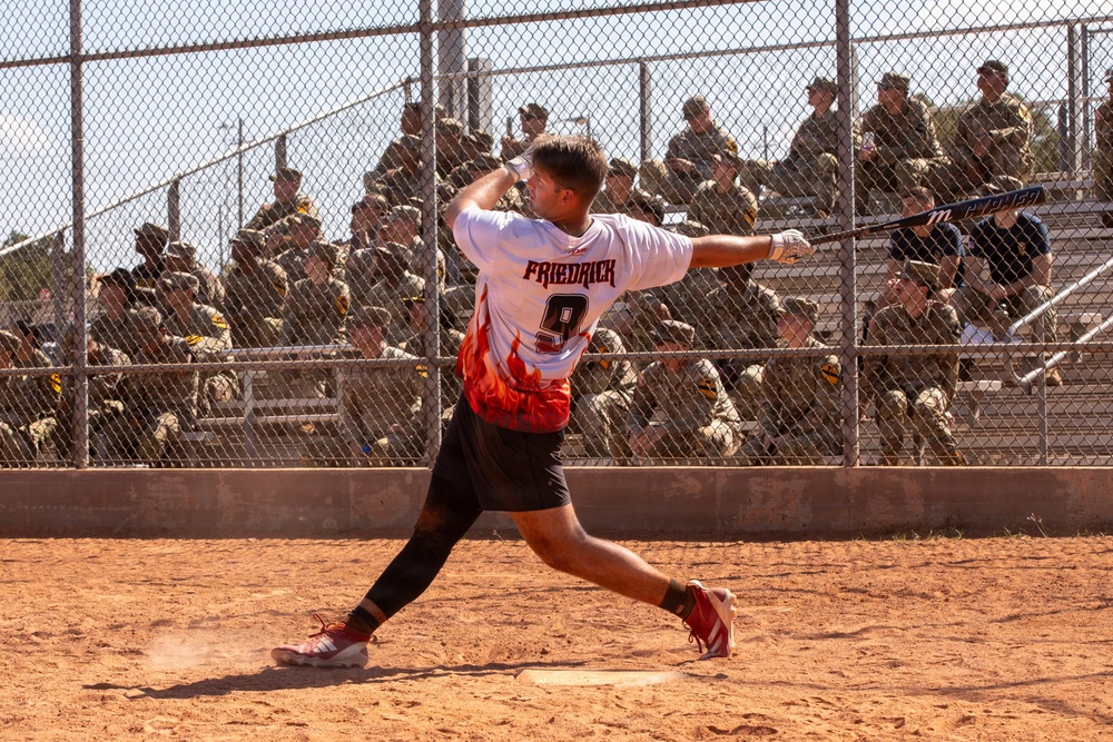 A baseball/softball player wearing jersey number 9 with the name 'FRIEDRICK' is captured mid-swing on a dirt infield. The player is wearing a white jersey with red flame graphics and black shorts. A large crowd of spectators dressed in military camouflage uniforms watches from bleachers behind a chain-link fence.