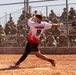 A baseball/softball player wearing jersey number 9 with the name 'FRIEDRICK' is captured mid-swing on a dirt infield. The player is wearing a white jersey with red flame graphics and black shorts. A large crowd of spectators dressed in military camouflage uniforms watches from bleachers behind a chain-link fence.