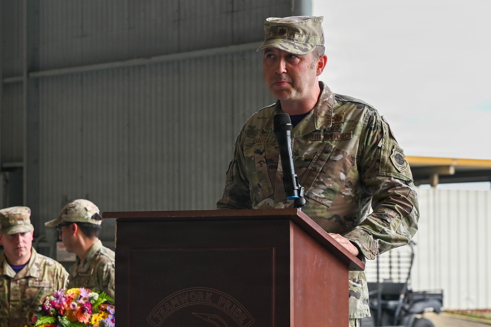 A U.S. Air Force officer in OCP (Operational Camouflage Pattern) uniform is speaking at a podium during what appears to be a formal military ceremony. The podium bears the inscription 'TEAMWORK PRIDE' and the speaker's uniform displays 'AIR FORCE' tape and various patches. Additional military personnel in similar uniforms are visible in the background.