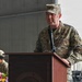 A U.S. Air Force officer in OCP (Operational Camouflage Pattern) uniform is speaking at a podium during what appears to be a formal military ceremony. The podium bears the inscription 'TEAMWORK PRIDE' and the speaker's uniform displays 'AIR FORCE' tape and various patches. Additional military personnel in similar uniforms are visible in the background.