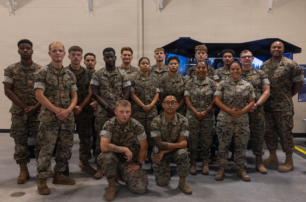 A group of approximately 20 military personnel in U.S. Marine Corps MARPAT (digital camouflage) uniforms pose for a formal group photograph inside what appears to be a training facility or simulator bay. The group is diverse in gender and ethnicity, arranged in two standing rows with two individuals kneeling in the front. A military simulator or training equipment is visible in the background.