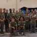 A group of approximately 20 military personnel in U.S. Marine Corps MARPAT (digital camouflage) uniforms pose for a formal group photograph inside what appears to be a training facility or simulator bay. The group is diverse in gender and ethnicity, arranged in two standing rows with two individuals kneeling in the front. A military simulator or training equipment is visible in the background.