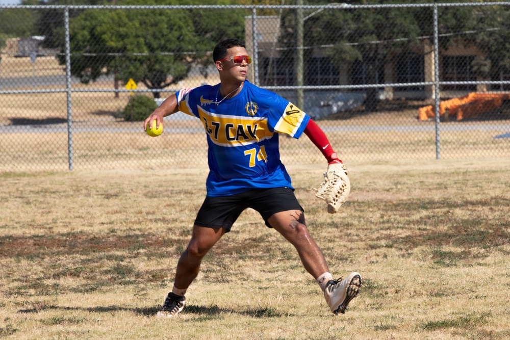 A softball player wearing a blue and gold '1-7 CAV' jersey with number 74 is in the process of throwing a yellow softball on a dry grass field. The player is wearing red sunglasses, a red arm sleeve, and holds a fielding glove in the opposite hand. The setting appears to be a recreational or military unit softball game based on the team name.