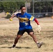 A softball player wearing a blue and gold '1-7 CAV' jersey with number 74 is in the process of throwing a yellow softball on a dry grass field. The player is wearing red sunglasses, a red arm sleeve, and holds a fielding glove in the opposite hand. The setting appears to be a recreational or military unit softball game based on the team name.