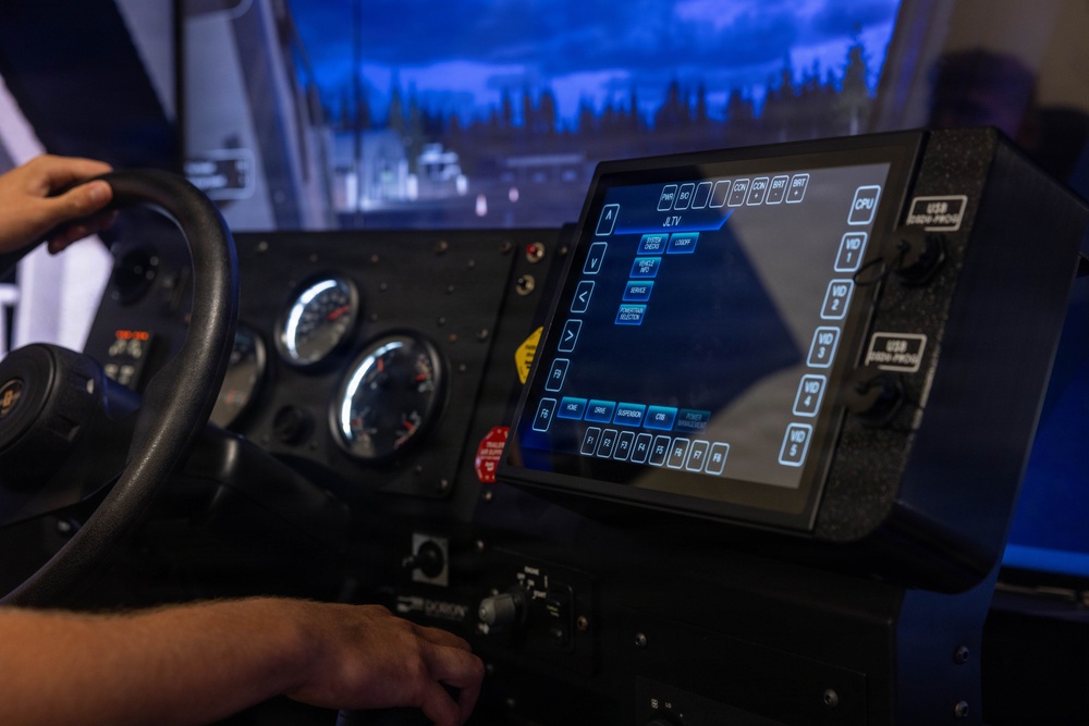 The image shows the interior of a military vehicle simulator, specifically a JLTV (Joint Light Tactical Vehicle) training simulator. A person's hands are visible on the steering wheel and control panel, interacting with a touchscreen display showing a menu interface. The setup includes analog gauges, control switches, and a projected simulation environment visible through the windshield.