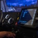 The image shows the interior of a military vehicle simulator, specifically a JLTV (Joint Light Tactical Vehicle) training simulator. A person's hands are visible on the steering wheel and control panel, interacting with a touchscreen display showing a menu interface. The setup includes analog gauges, control switches, and a projected simulation environment visible through the windshield.