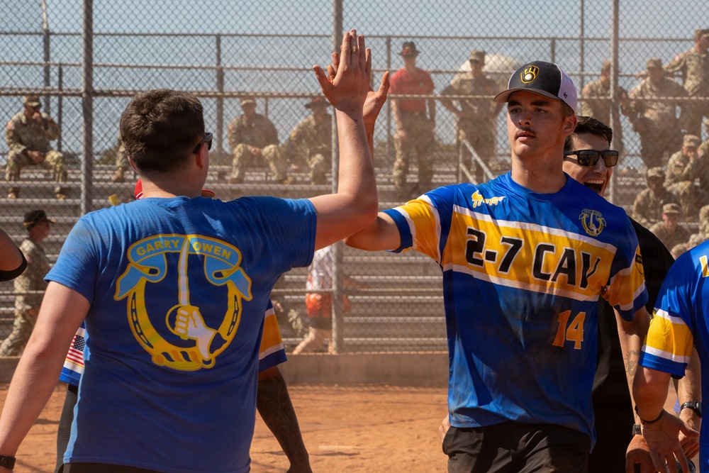 Two individuals are exchanging a high-five on a dirt softball/baseball field during what appears to be a military unit sporting event. One person wears a blue '2-7 CAV' (2nd Squadron, 7th Cavalry) jersey with number 14, while the other wears a blue 'Garry Owen' t-shirt referencing the 7th Cavalry Regiment's traditional march. A large crowd of military personnel in uniform watches from bleachers in the background.