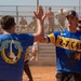 Two individuals are exchanging a high-five on a dirt softball/baseball field during what appears to be a military unit sporting event. One person wears a blue '2-7 CAV' (2nd Squadron, 7th Cavalry) jersey with number 14, while the other wears a blue 'Garry Owen' t-shirt referencing the 7th Cavalry Regiment's traditional march. A large crowd of military personnel in uniform watches from bleachers in the background.
