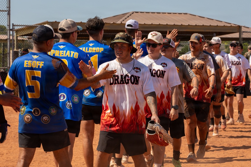 Two softball teams are engaged in a post-game handshake line on a dirt baseball/softball field. One team wears blue jerseys with military branch insignia patches and player names (including 'ESOFF #5', 'UPREIATO', 'WALDROP'), while the opposing team wears white and red flame-design jerseys labeled 'DRAGON'. The setting appears to be a military recreational softball event based on the uniform patches representing Army, Navy, Air Force, and other branches.