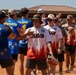 Two softball teams are engaged in a post-game handshake line on a dirt baseball/softball field. One team wears blue jerseys with military branch insignia patches and player names (including 'ESOFF #5', 'UPREIATO', 'WALDROP'), while the opposing team wears white and red flame-design jerseys labeled 'DRAGON'. The setting appears to be a military recreational softball event based on the uniform patches representing Army, Navy, Air Force, and other branches.
