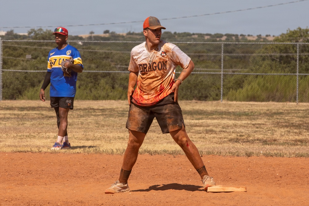 Two players are on a dirt softball/baseball field during an active game. The foreground player, wearing a 'Dragon' jersey with flame graphics, stands on a base in a ready stance with visibly muddy and dirty clothing suggesting heavy play. A second player in a blue and yellow jersey stands in the background holding a glove.