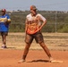 Two players are on a dirt softball/baseball field during an active game. The foreground player, wearing a 'Dragon' jersey with flame graphics, stands on a base in a ready stance with visibly muddy and dirty clothing suggesting heavy play. A second player in a blue and yellow jersey stands in the background holding a glove.