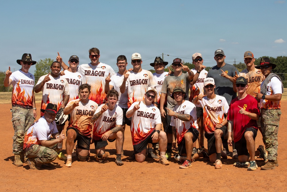 A group of approximately 20 men pose for a team photo on a dirt softball/baseball field. Most members are wearing matching white 'DRAGON' jerseys with flame designs at the bottom, while some wear military camouflage uniforms. Several individuals are holding up index fingers in a 'number one' gesture, suggesting a victory or championship celebration.