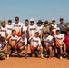 A group of approximately 20 men pose for a team photo on a dirt softball/baseball field. Most members are wearing matching white 'DRAGON' jerseys with flame designs at the bottom, while some wear military camouflage uniforms. Several individuals are holding up index fingers in a 'number one' gesture, suggesting a victory or championship celebration.
