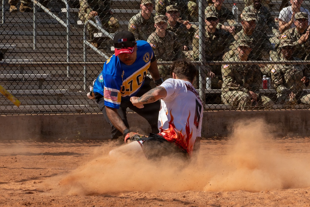 A dynamic softball or baseball game is captured mid-action, showing a player in a white uniform with flame graphics sliding into a base while a fielder in a blue jersey attempts a tag. A large cloud of red-brown dirt dust has been kicked up by the slide. The game is being watched by a crowd of military personnel in camouflage uniforms seated in bleachers behind a chain-link fence.