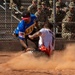 A dynamic softball or baseball game is captured mid-action, showing a player in a white uniform with flame graphics sliding into a base while a fielder in a blue jersey attempts a tag. A large cloud of red-brown dirt dust has been kicked up by the slide. The game is being watched by a crowd of military personnel in camouflage uniforms seated in bleachers behind a chain-link fence.