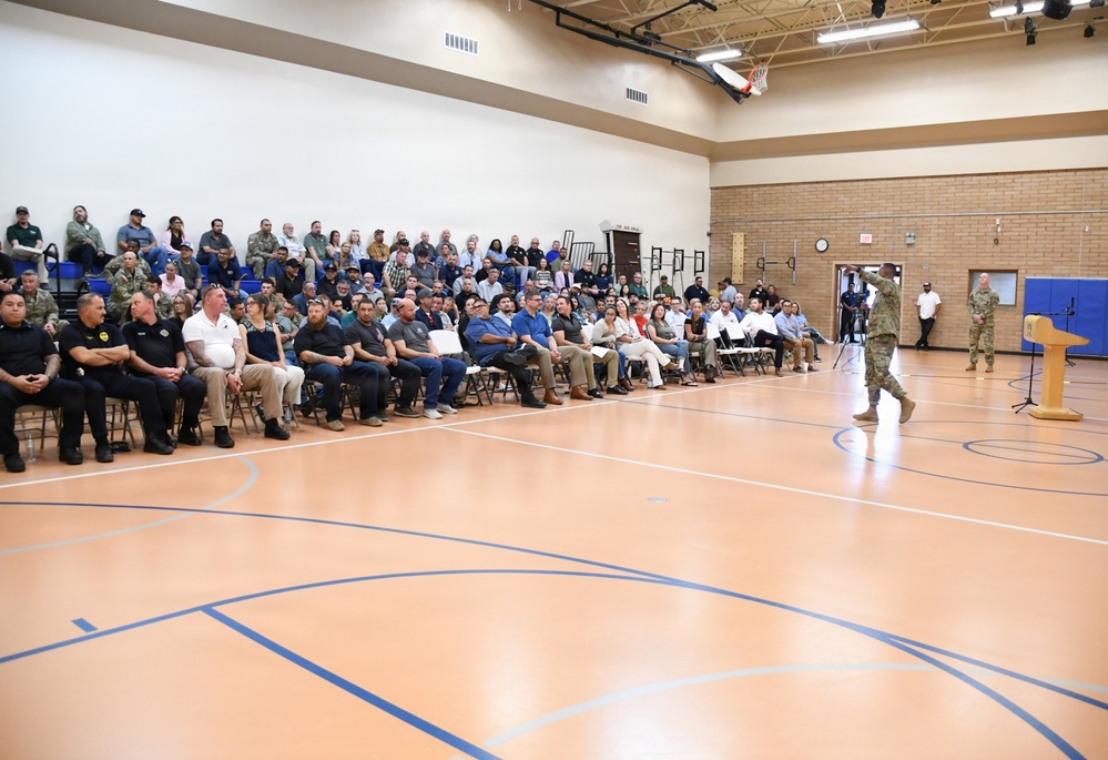 A large gathering is taking place inside a military or government gymnasium/multipurpose facility. A person in military uniform is addressing a sizable audience seated in chairs and on bleachers. A podium is visible on the right side of the court, suggesting a formal briefing or town hall-style event.