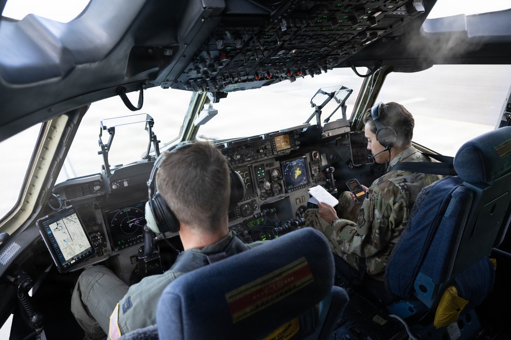 Two military aircrew members are seated in the cockpit of a large military transport aircraft, likely a C-17 Globemaster III or similar aircraft. Both personnel are wearing flight headsets and military uniforms — one in an olive flight suit and one in OCP (Operational Camouflage Pattern) uniform. The aircraft appears to be on the ground during pre-flight or ground operations.