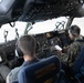 Two military aircrew members are seated in the cockpit of a large military transport aircraft, likely a C-17 Globemaster III or similar aircraft. Both personnel are wearing flight headsets and military uniforms — one in an olive flight suit and one in OCP (Operational Camouflage Pattern) uniform. The aircraft appears to be on the ground during pre-flight or ground operations.