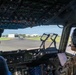 A military aircrew member wearing a Bose headset and camouflage uniform is seated in the cockpit of a large military transport aircraft, viewed from behind. The cockpit features an extensive array of overhead panels, digital displays, and flight instruments. Through the windscreen, a military airfield is visible with what appear to be fighter jets taxiing on the tarmac and hangars in the background.