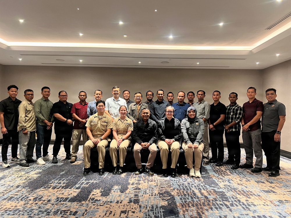 A group photograph of approximately 25 individuals posed together in what appears to be a hotel conference or banquet room. The group includes both military personnel in uniform and civilians in casual or business-casual attire. Two individuals in the front row are wearing what appear to be U.S. Navy uniforms, suggesting a multinational or joint military-civilian gathering.