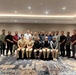 A group photograph of approximately 25 individuals posed together in what appears to be a hotel conference or banquet room. The group includes both military personnel in uniform and civilians in casual or business-casual attire. Two individuals in the front row are wearing what appear to be U.S. Navy uniforms, suggesting a multinational or joint military-civilian gathering.