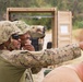The image shows military personnel in camouflage uniforms and helmets engaged in what appears to be a training exercise at an outdoor range or tactical training area. One service member is pointing forward, apparently directing or instructing others, while additional personnel are visible in the background near a tan shipping container. The setting includes natural vegetation and appears to be a controlled military training environment.