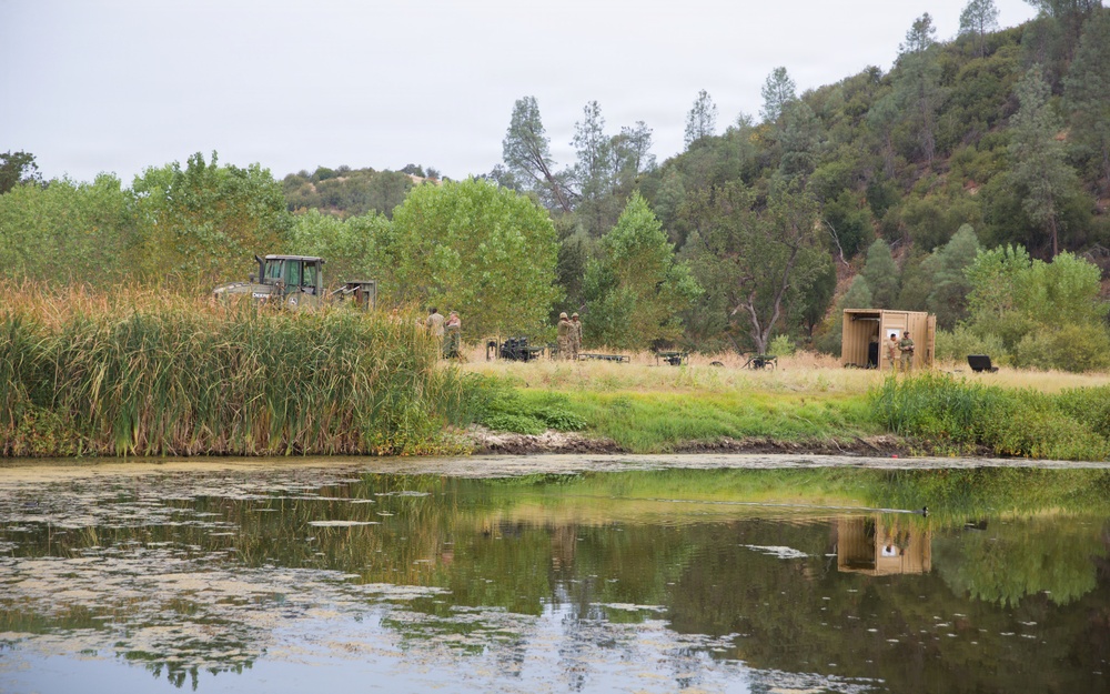 The image shows a military field operation set up along the bank of a calm reflective pond or river, surrounded by dense vegetation and wooded hills. Several personnel in military camouflage uniforms are visible near equipment, a tractor, and a tan/wooden modular structure or shelter. The scene appears to be a training exercise or field deployment in a rural, semi-arid landscape.