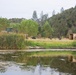 The image shows a military field operation set up along the bank of a calm reflective pond or river, surrounded by dense vegetation and wooded hills. Several personnel in military camouflage uniforms are visible near equipment, a tractor, and a tan/wooden modular structure or shelter. The scene appears to be a training exercise or field deployment in a rural, semi-arid landscape.