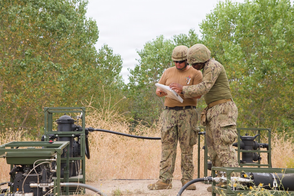Two military personnel in camouflage uniforms and combat helmets are reviewing documents together in a field environment. They are surrounded by green military pump or generator equipment connected by hoses and pipes. The setting appears to be a training exercise or field operation in an arid, semi-vegetated outdoor area.