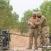 Two military personnel in camouflage uniforms and combat helmets are reviewing documents together in a field environment. They are surrounded by green military pump or generator equipment connected by hoses and pipes. The setting appears to be a training exercise or field operation in an arid, semi-vegetated outdoor area.