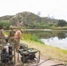 Three military personnel in mixed uniforms are gathered near a pond or small lake, reviewing documents or equipment. Multiple military-green water purification or pumping units are staged along the shoreline, connected by hoses. The setting appears to be a training or field operation site in a semi-arid, hilly landscape.